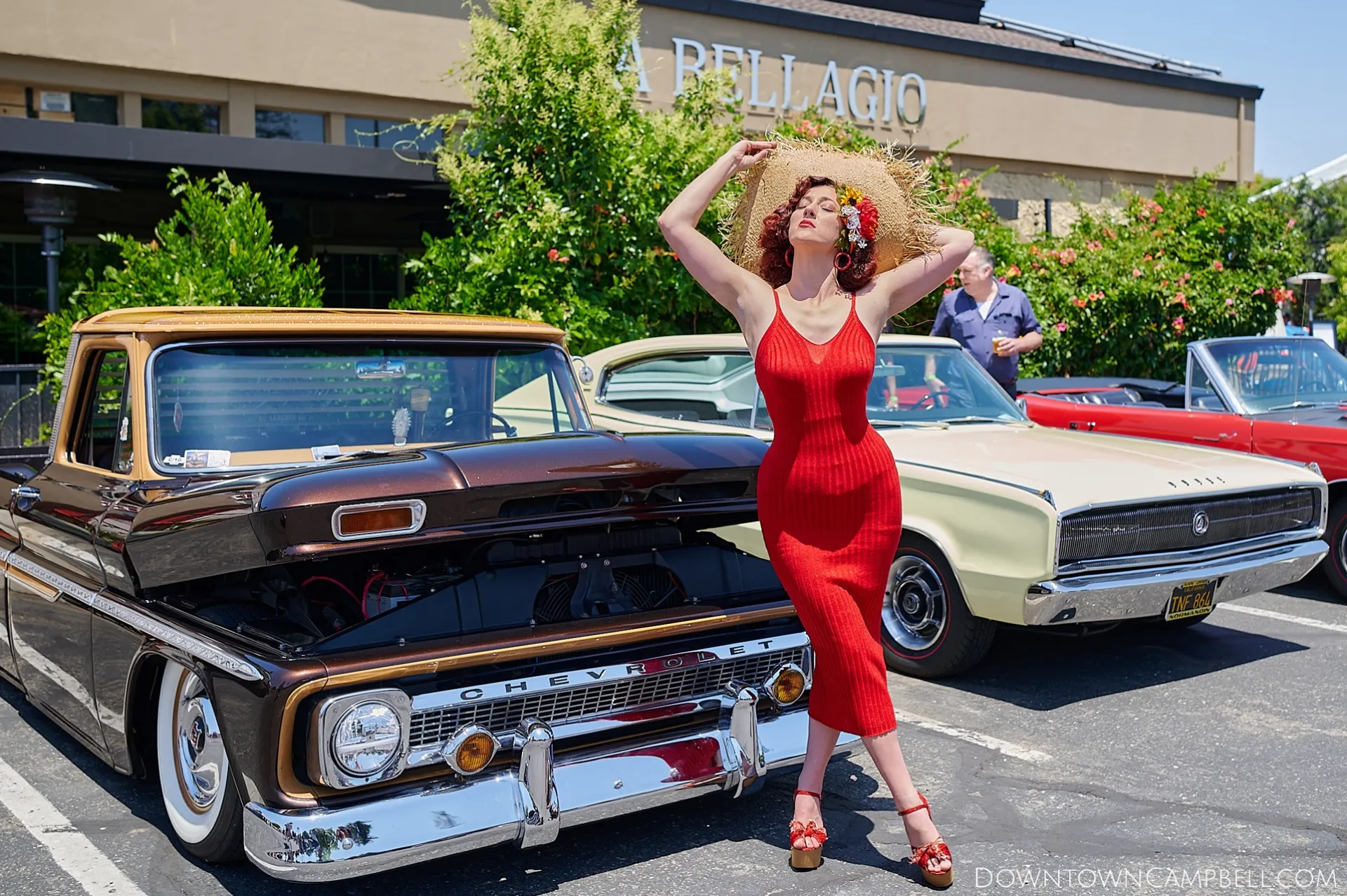 A voluptuous woman in red spaghetti strap dress poses next to a classic Ford truck converted into a low rider with an exquisite custom etched trim. She's looking up toward the sky as she adjusts her straw hat.