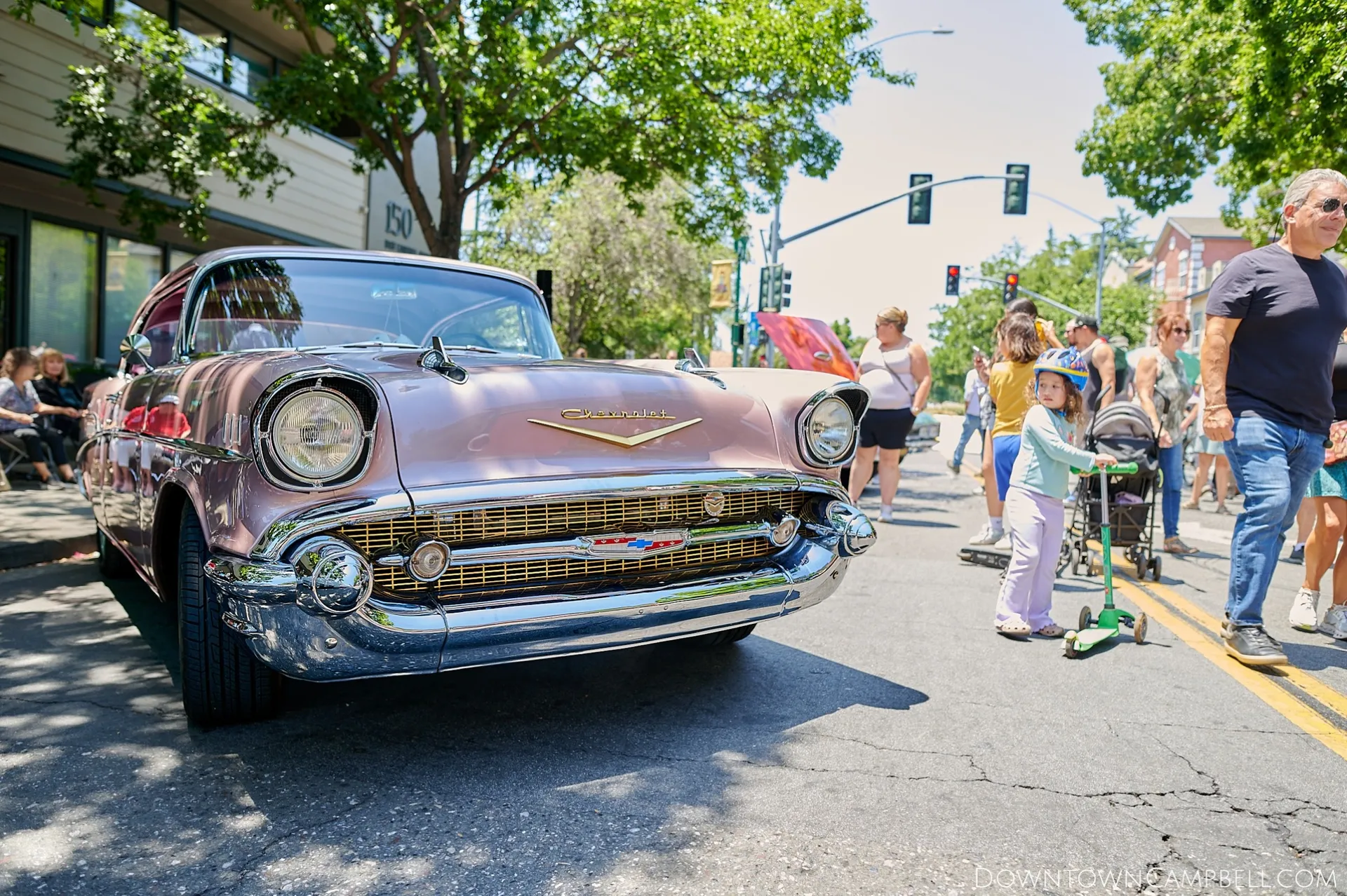A close-up look at the front of a rose-gold colored Chevy Bel-Air with gold accents. A child on a foot scooter pedals past the classic vehicle as onlookers admire the perfectly polished car.