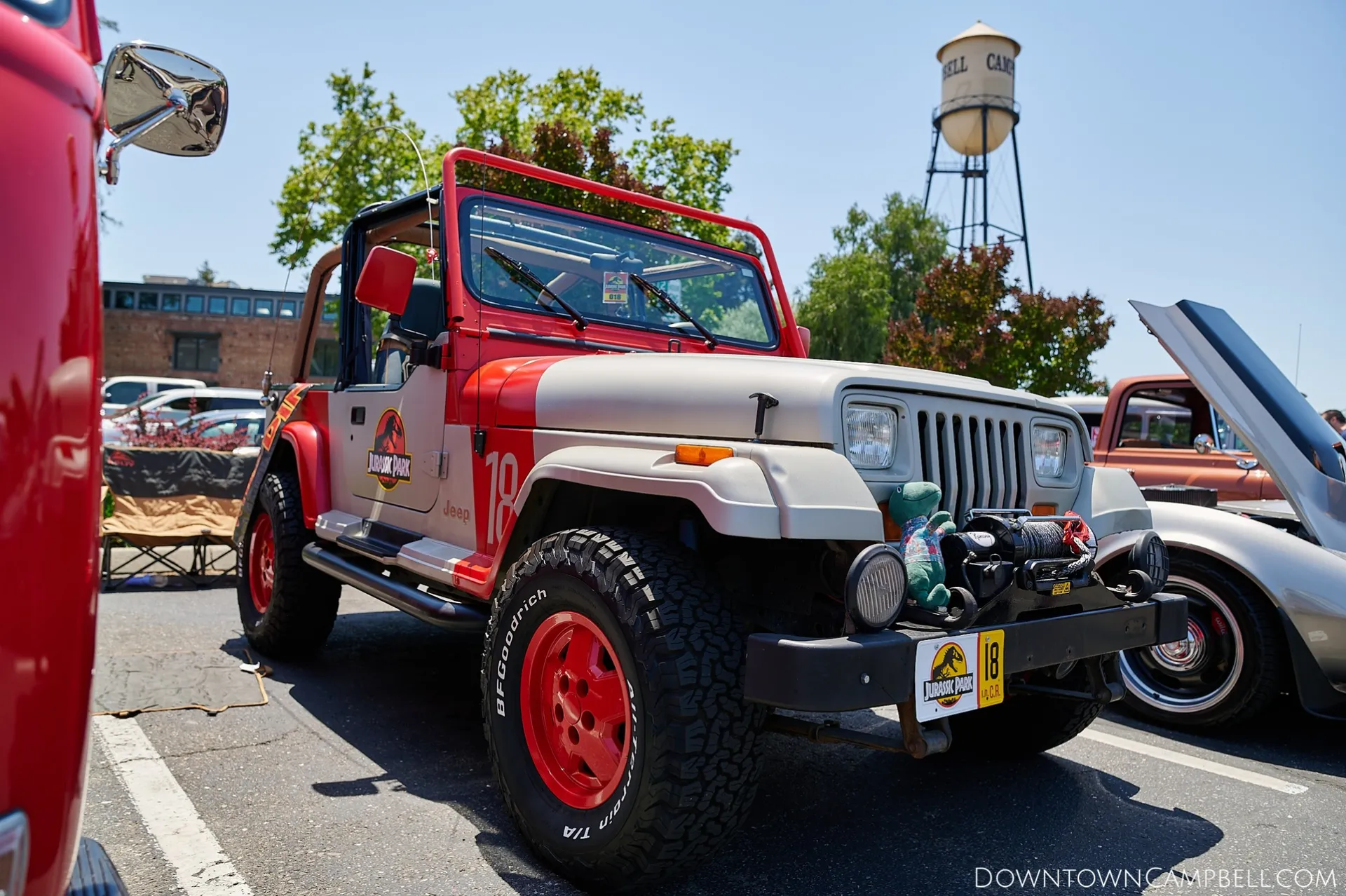 An offroad 4x4 jeep with decorated to look exactly like a jungle vehicle from the movie Jurrasic Park. The jeep is tan and red all over with very shiny black offroad tires. On the door of the jeep is the Jurrasic Park logo.